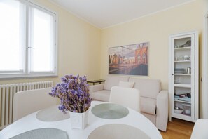 Un salón con un sofá blanco, una mesa de comedor con individuales, un jarrón de flores, un cuadro en la pared y un aparador con vajilla.