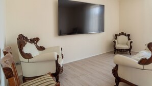 Una habitación con dos sillones ornamentados, un televisor de pantalla plana grande en la pared y un suelo de madera en tono claro.