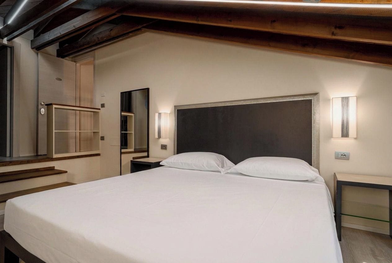 A modern bedroom with a large bed, bedside tables, a mirror, and built-in lighting.