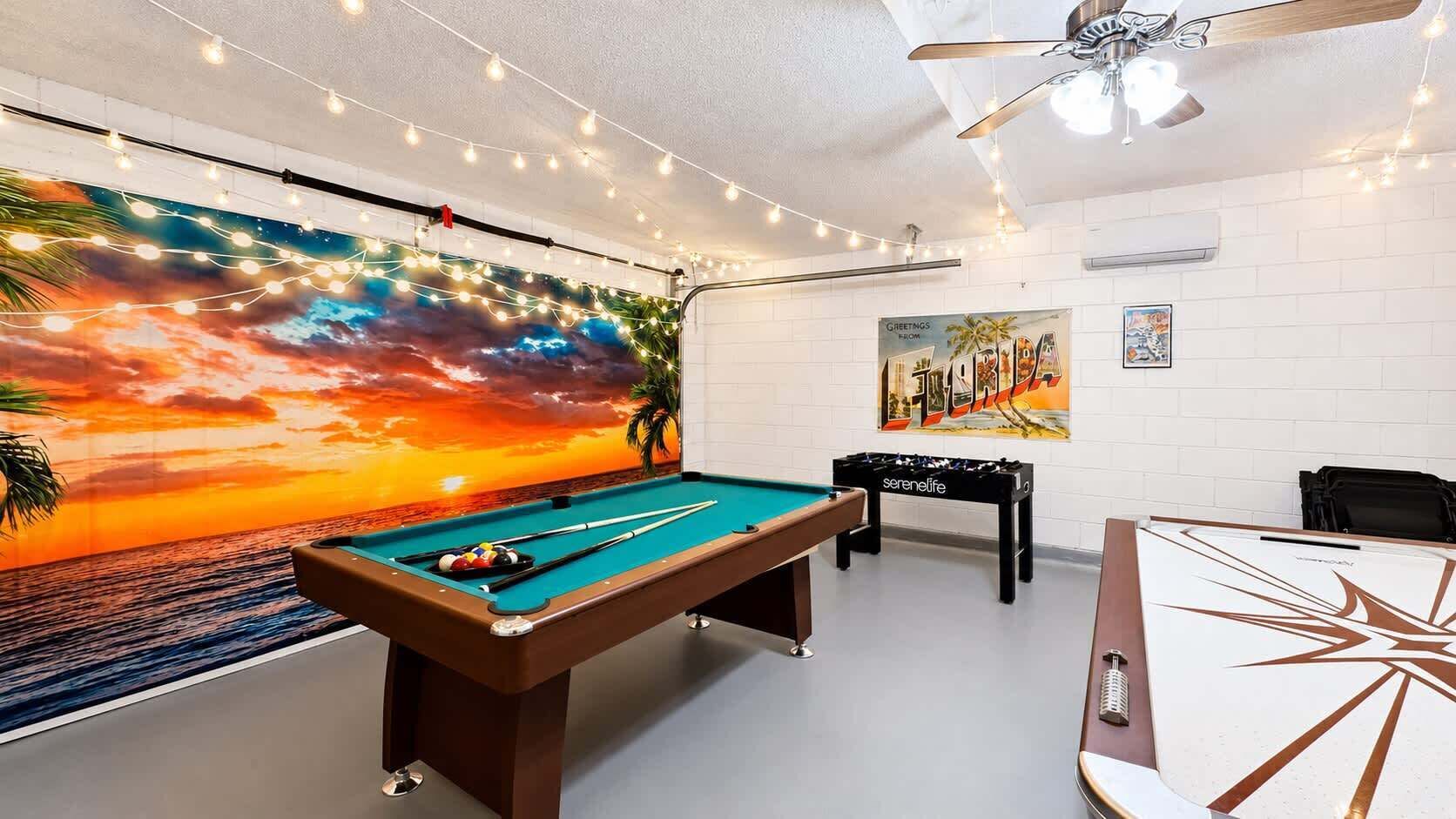 Ein Billardtisch mit Sonnenuntergangsmotiv, ein Tischfußballspiel und ein Deckenventilator in einem Raum mit weißen Wänden und Lichterketten.