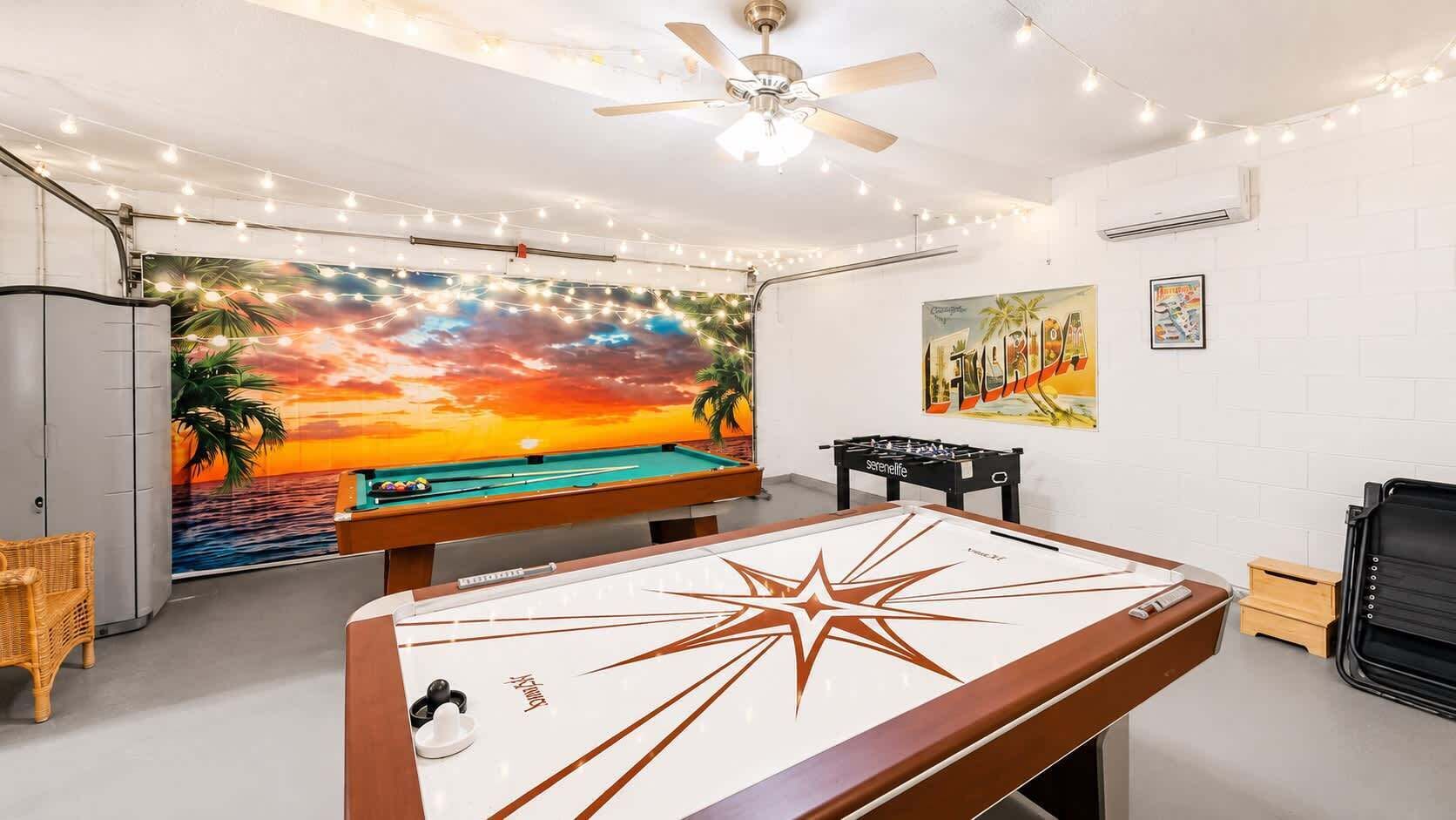 Une table de billard ornée d’un motif en étoile, une table de baby-foot, un ventilateur de plafond, des guirlandes lumineuses et une fresque représentant un coucher de soleil.