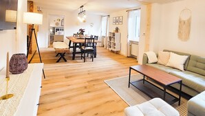 Een ruime woonkamer met houten vloer, een eettafel met stoelen, een bank en een salontafel. Er is een lamp en een wandlamp.