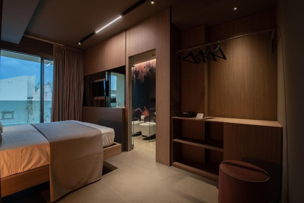 A modern hotel room with a bed, a walk-in closet, and a bathroom visible through a glass door.