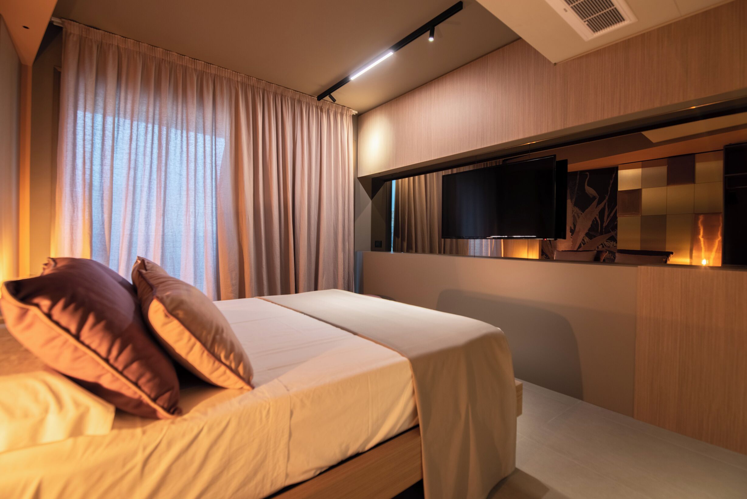 A modern hotel room with a bed, curtains, a mounted TV, and a wooden wall panel.