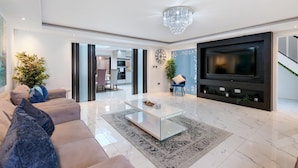 A modern living room with a glass coffee table, a large flat-screen TV, a chandelier, and a sofa with decorative pillows.
