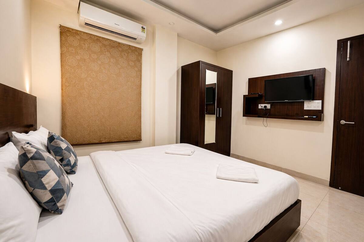 A hotel room with a bed, pillows, a TV mounted on the wall, a wardrobe, and an air conditioning unit.