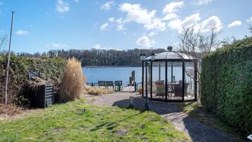 A gazebo with a glass enclosure overlooking a lake, surrounded by a grassy area and a hedge.