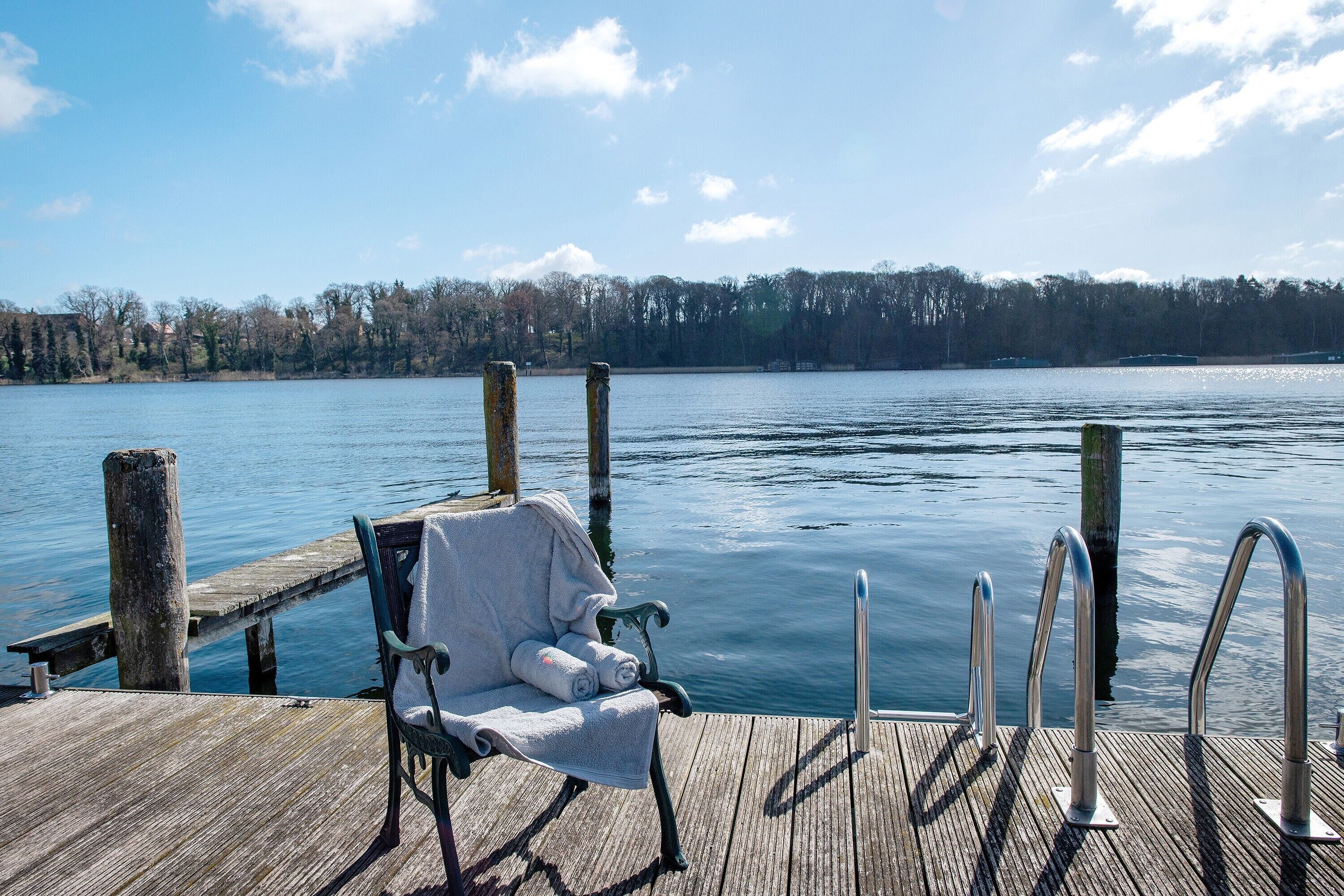 Eine Holzsteganlage mit Stuhl und Handtuch, mit Blick auf einen stillen See mit Bäumen und blauem Himmel.