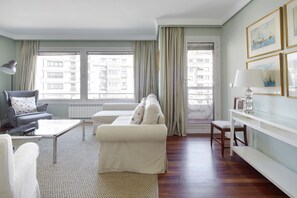 A living room with a sofa, armchair, and a coffee table. There are curtains on the windows, and a lamp on a side table. - Koxka - Basque Stay (San Sebastián)