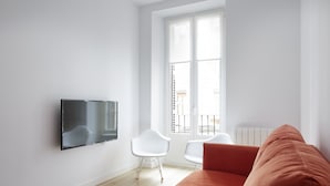 A modern living room with a flat-screen TV mounted on the wall, two white chairs, a large orange sofa, and a window with a view of buildings. - Etxaide - Basque Stay (San Sebastián)