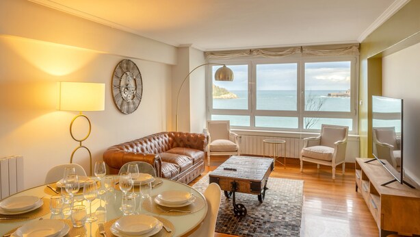 A living room with a leather sofa, dining table set for six, a coffee table, and a view of the sea through large windows. - Maskortxo - Basque Stay (San Sebastián)