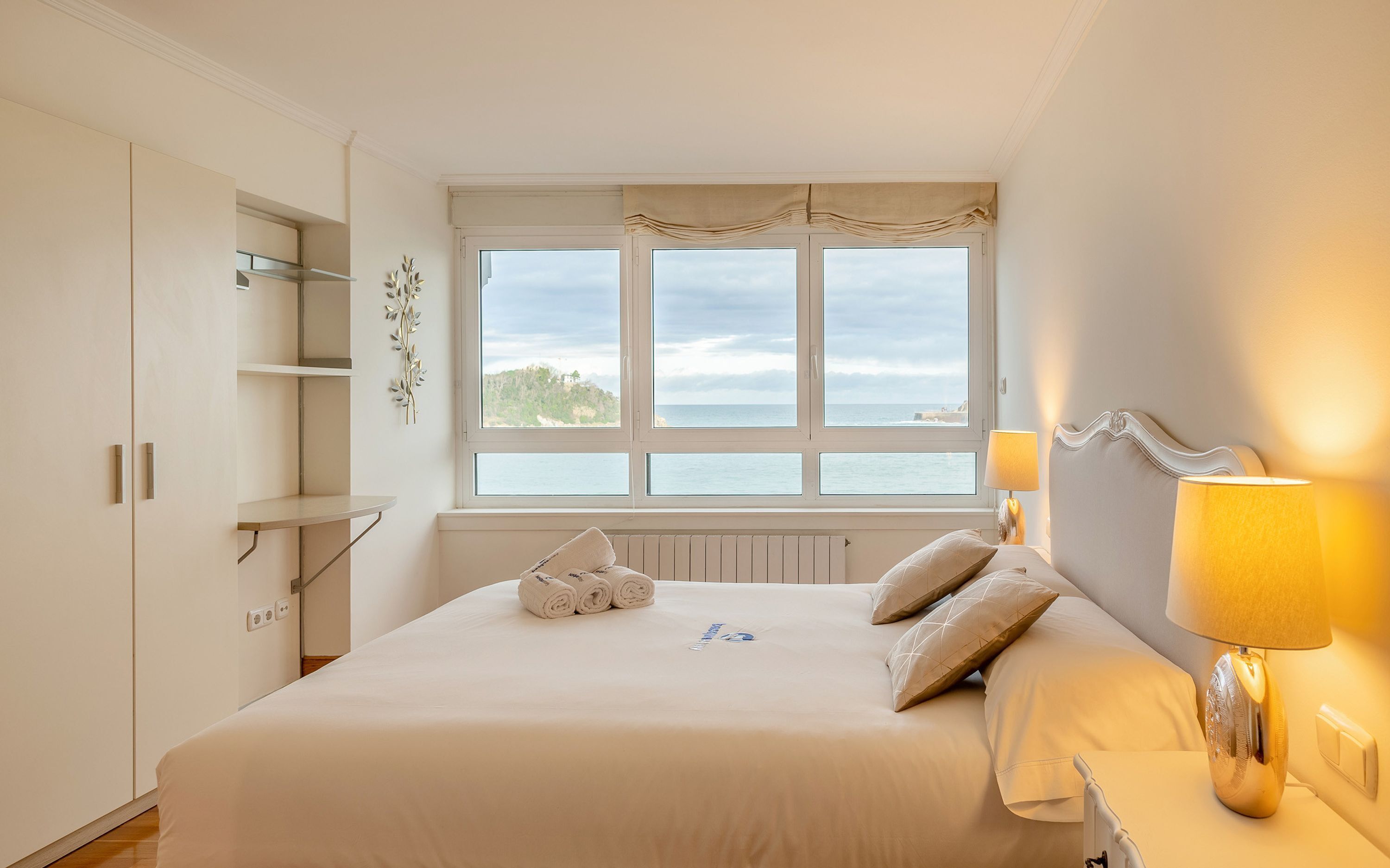 A bedroom with a large bed, a window with a view of the sea, and a bedside lamp.