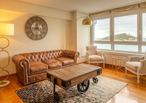 Un salon avec un canapé en cuir marron, une table basse en bois sur roulettes, un fauteuil blanc, une lampe sur pied et une grande horloge au mur.