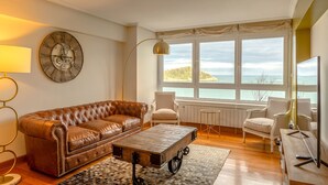 A living room with a brown leather sofa, a wooden coffee table with wheels, a floor lamp, a clock, and a view of the sea through large windows. - Maskortxo - Basque Stay (San Sebastián)