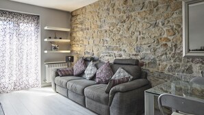 A modern living room with a stone wall, grey sofa, patterned cushions, and a glass table. - Ordulari - Basque Stay (San Sebastián)