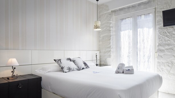 A modern bedroom with a large bed, white bedding, a nightstand with a lamp, and a stone wall. - Ordulari - Basque Stay (San Sebastián)