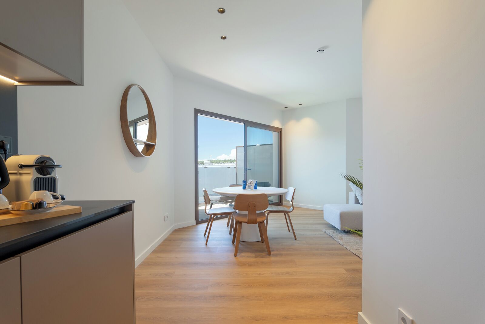 A modern kitchen with a coffee machine, a dining area with a round table and chairs, and a large sliding glass door leading to a balcony.