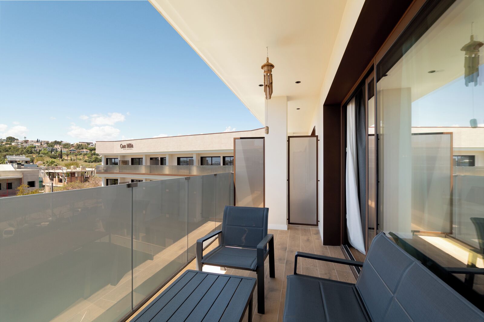Un balcón con barandillas de vidrio, dos sillas, una mesa y vistas de edificios al fondo.