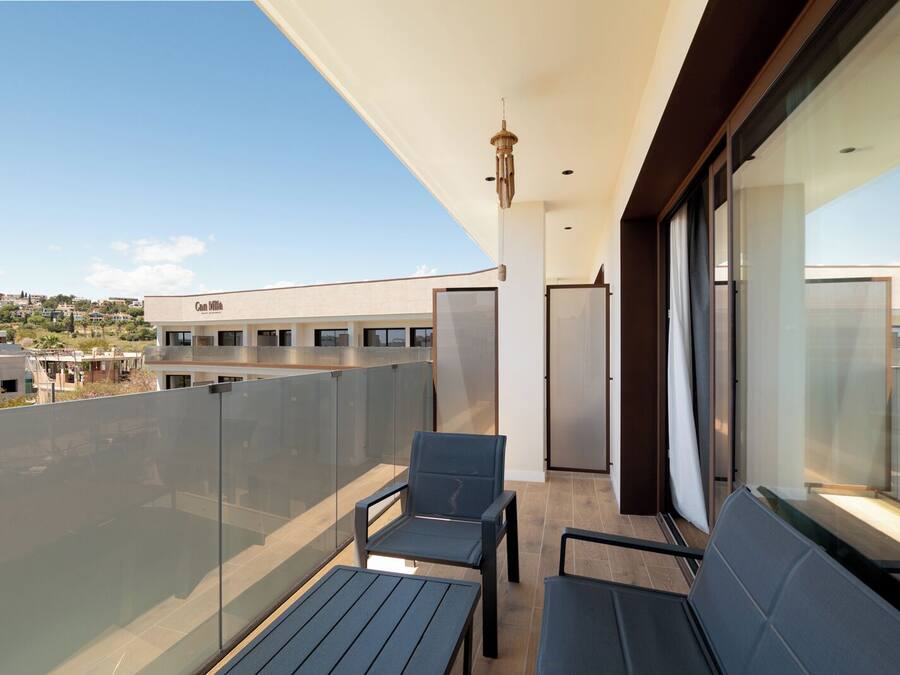 Un balcón con barandillas de vidrio, dos sillas, una mesa y vistas de edificios al fondo.