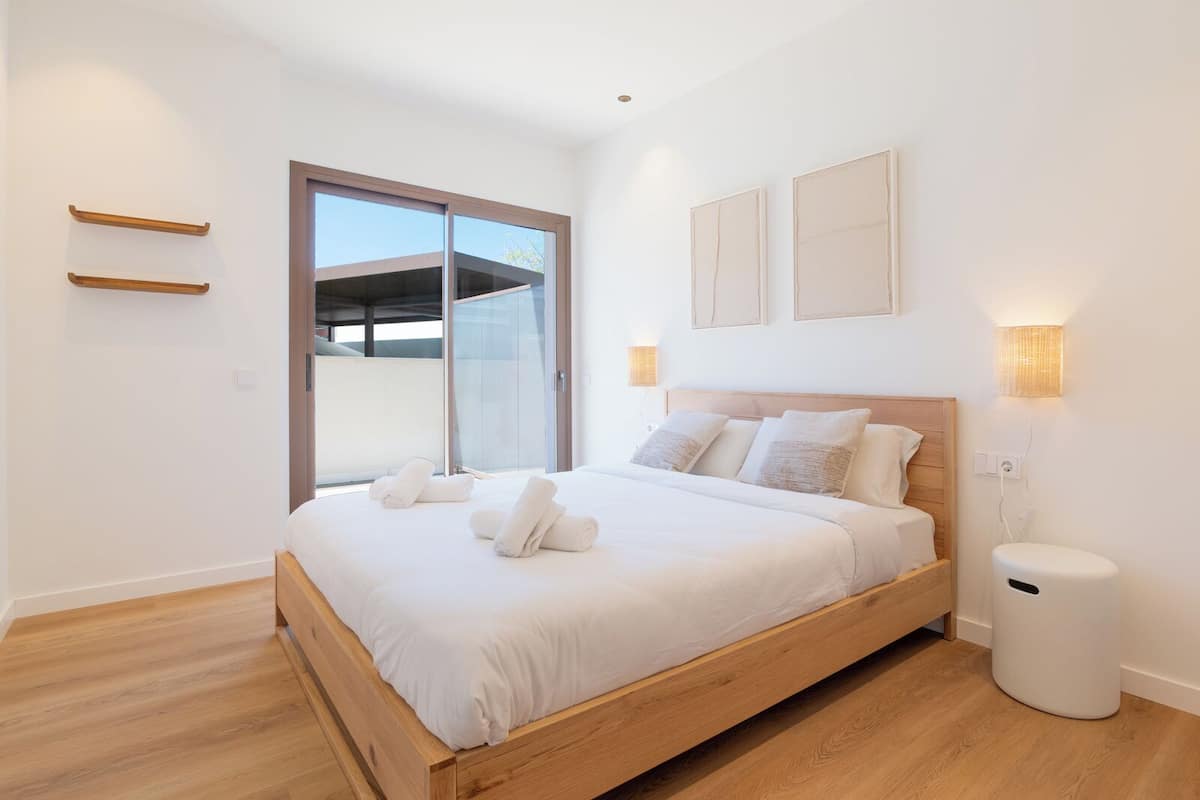 Un dormitorio con una cama de madera, una puerta corrediza de vidrio, un cesto de basura blanco y dos lámparas fijadas a la pared.