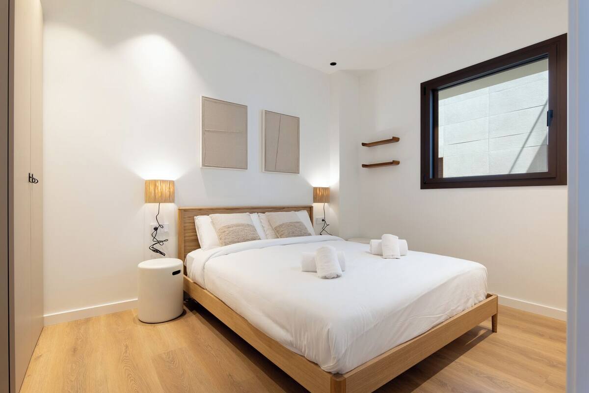 Un dormitorio ordenado con una cama de madera, lámparas de noche, una mesita de noche pequeña y una ventana con vistas.