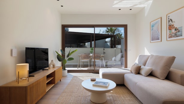 Ein modernes Wohnzimmer mit Flachbildfernseher, einem beigefarbenen Sofa, einem Couchtisch und einer Glastür, die zu einem Außenbereich mit Esstisch und Stühlen führt.
