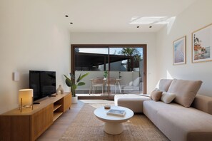 Ein modernes Wohnzimmer mit Flachbildfernseher, einem beigefarbenen Sofa, einem Couchtisch und einer Glastür, die zu einem Außenbereich mit Esstisch und Stühlen führt.