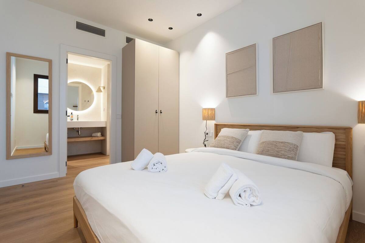 Una habitación de hotel moderna con una cama grande, un baño con espejo y lavamanos, y un armario.