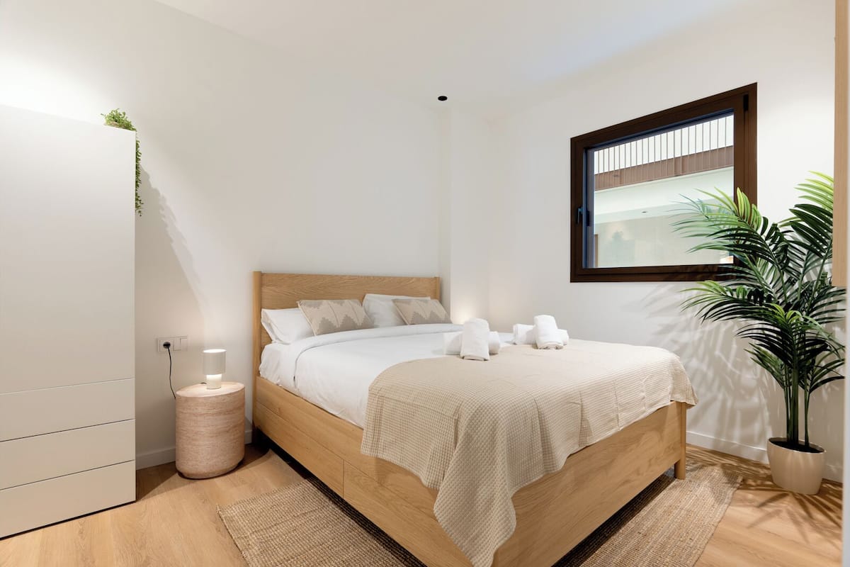 Un dormitorio moderno con cama de madera, lámparas de noche, una planta en maceta y un ventanal.