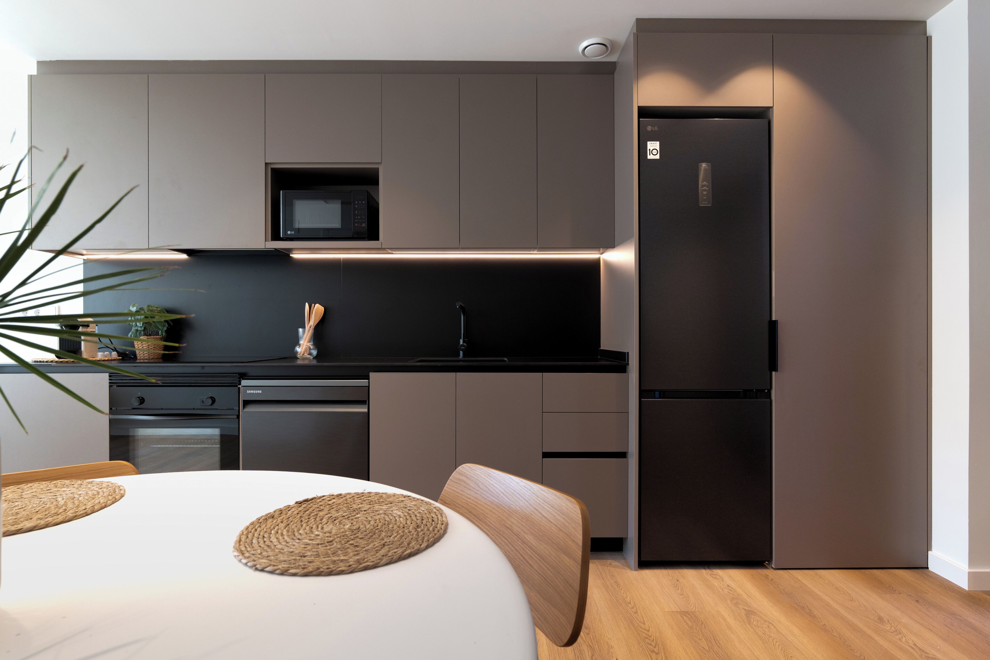 A modern kitchen with a built-in microwave, a stainless steel refrigerator, and a dining area with a white table and woven placemats.