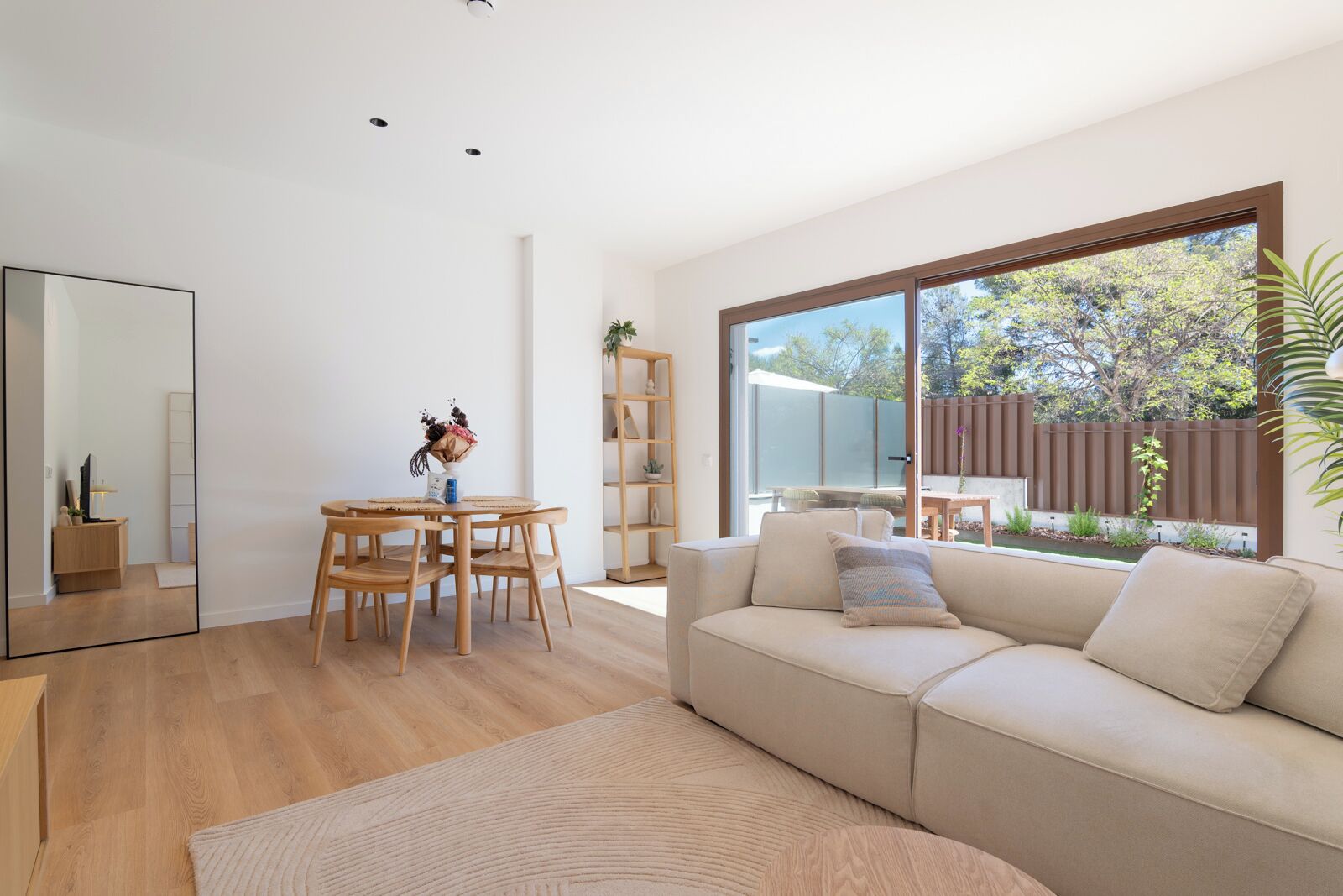 A modern living room with a sofa, a dining table with chairs, a large window, and a bookshelf.