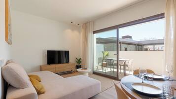 Una habitación de hotel moderna con sofá, televisor, mesa de comedor y balcón con mobiliario exterior.