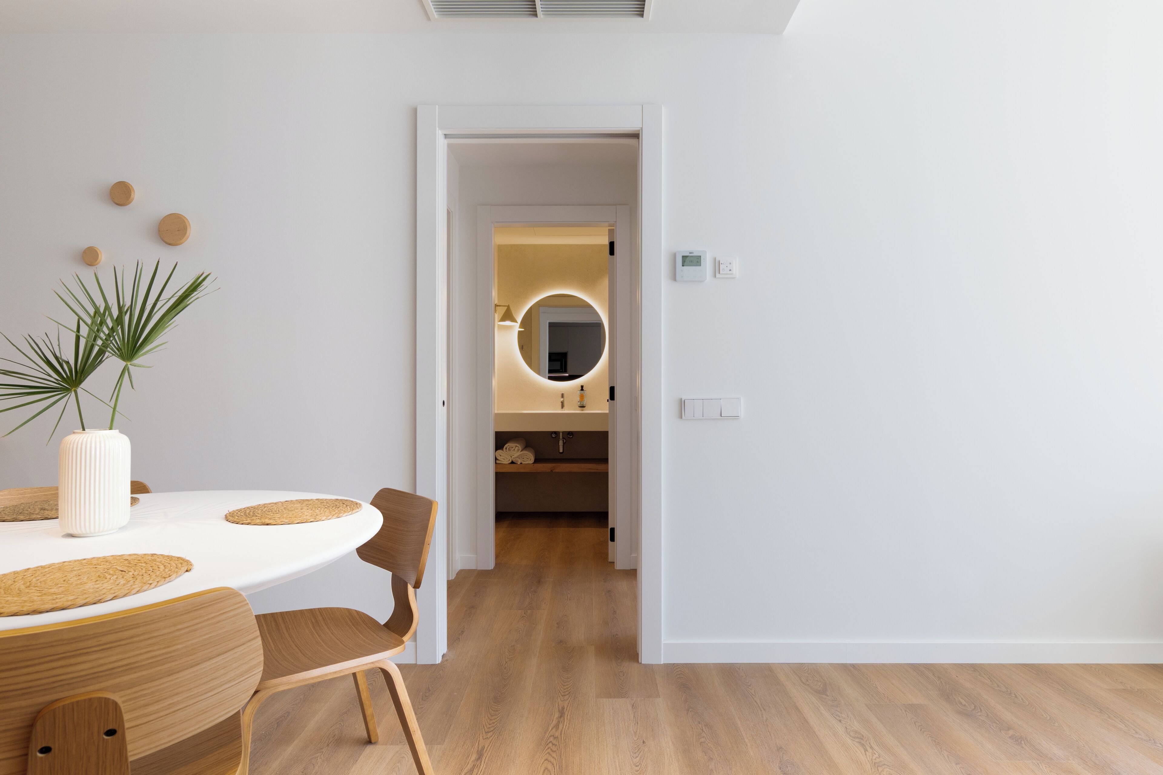 A modern interior with a dining table, chairs, a potted plant, and a hallway leading to a bathroom with a round mirror.