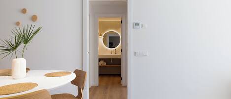 A modern interior with a dining table, chairs, a potted plant, and a hallway leading to a bathroom with a round mirror.