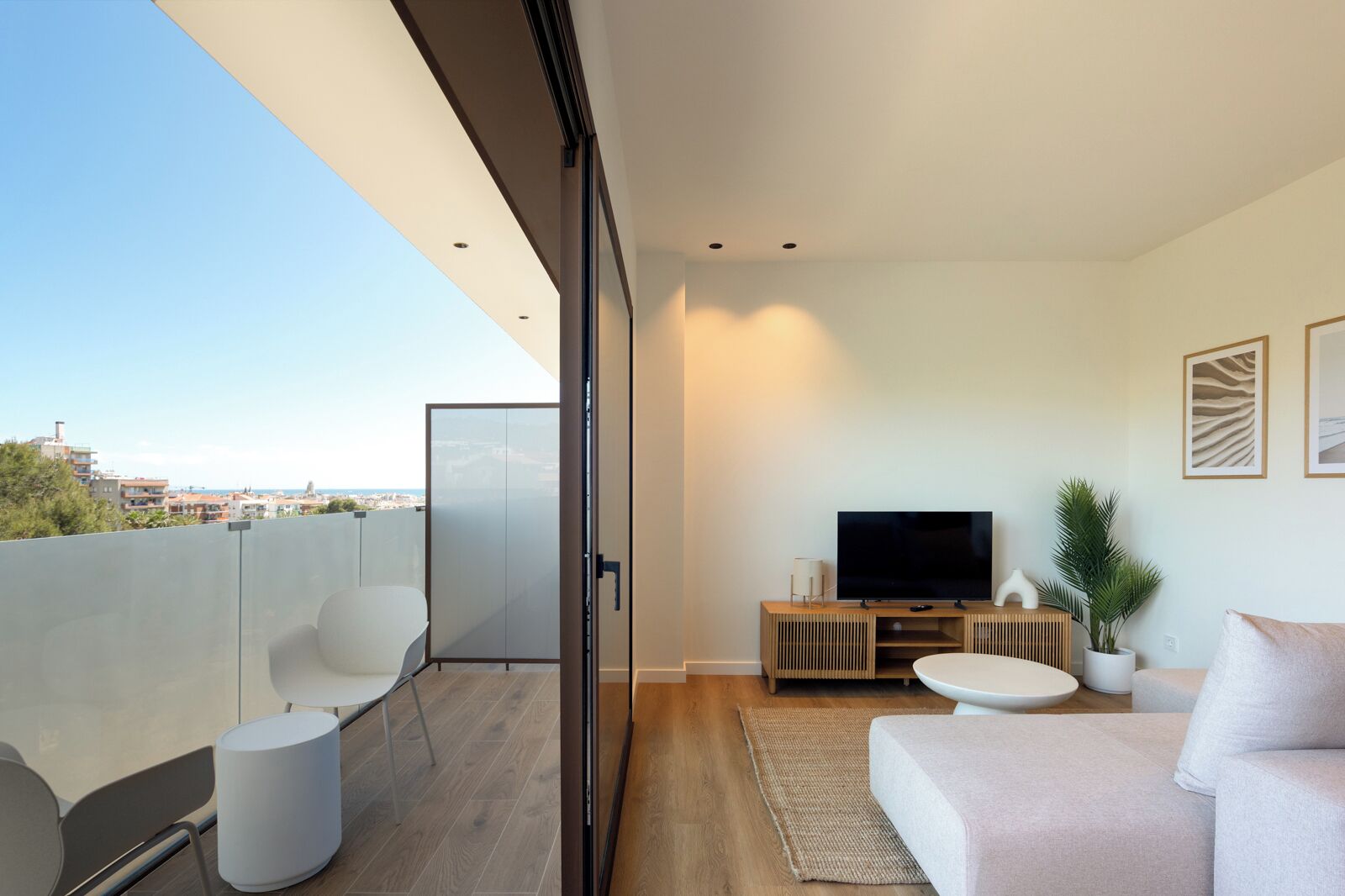 A modern living room with a TV, sofa, and chairs, a balcony with a view of the city, and a sliding glass door.