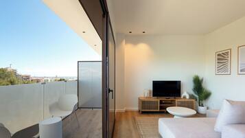 A modern living room with a TV, sofa, and chairs, a balcony with a view of the city, and a sliding glass door.