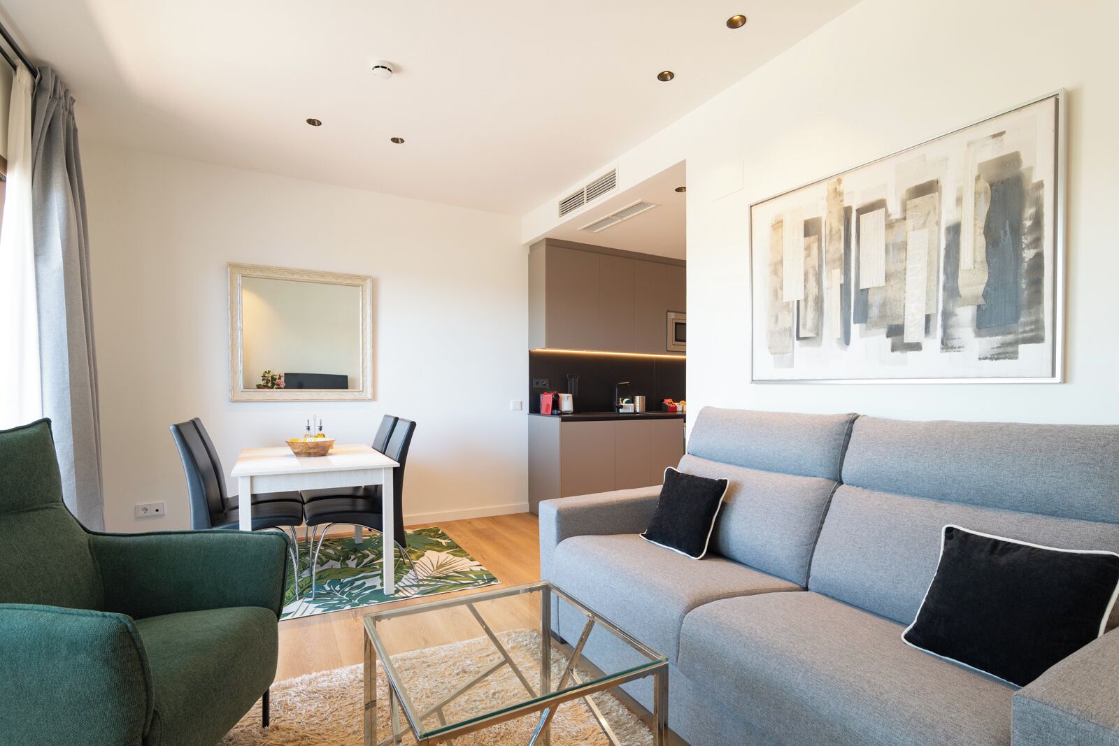 Un salon moderne avec un canapé gris, un fauteuil vert, une table à manger avec des chaises, et une cuisine en arrière-plan.
