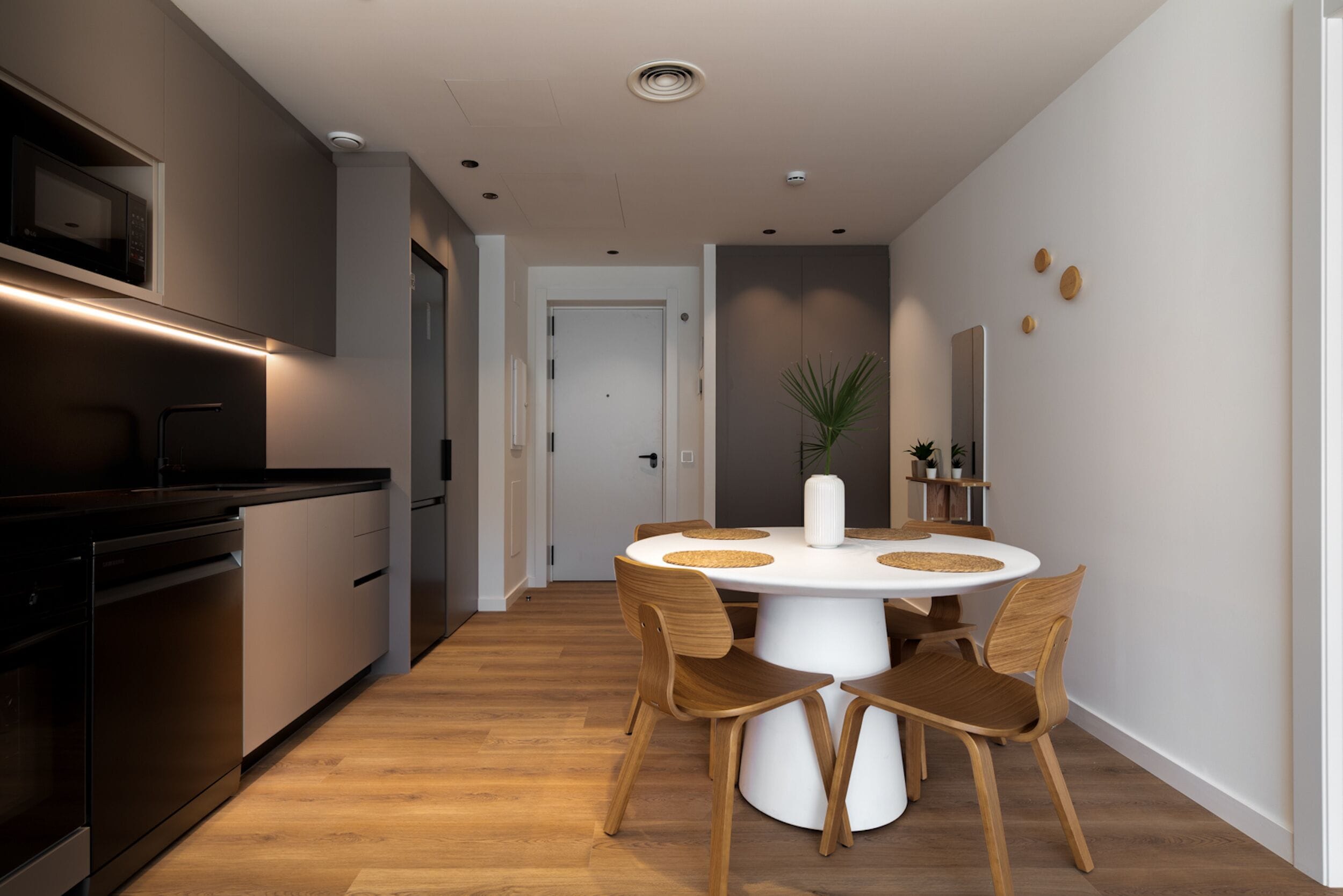 A modern kitchen with built-in appliances, a dining area with a round table and wooden chairs, and a hallway leading to a door.