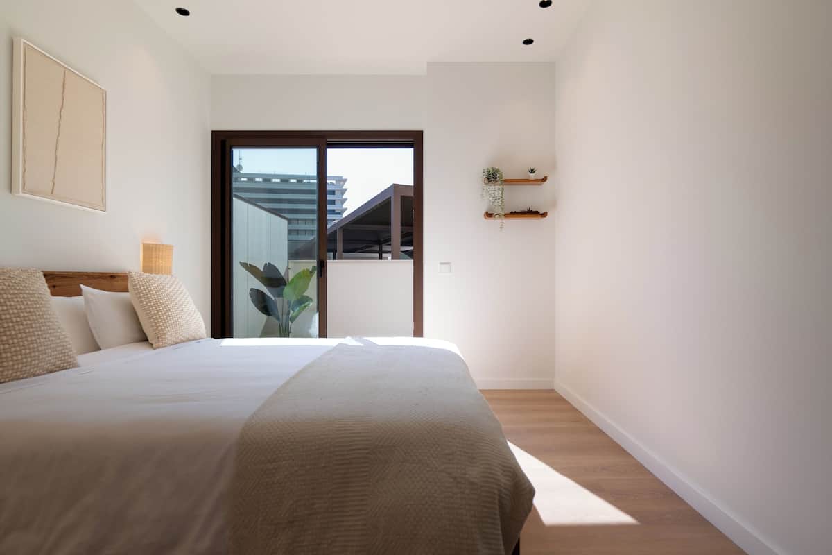 Un dormitorio con cama, un ventanal amplio, una planta y una estantería con plantas.