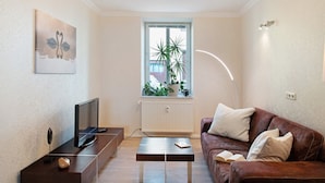 A living room with a TV, a coffee table, a sofa, a window with plants, and a painting on the wall. - Apartment 'Fewo Schwan' with Wi-Fi (Malchow)