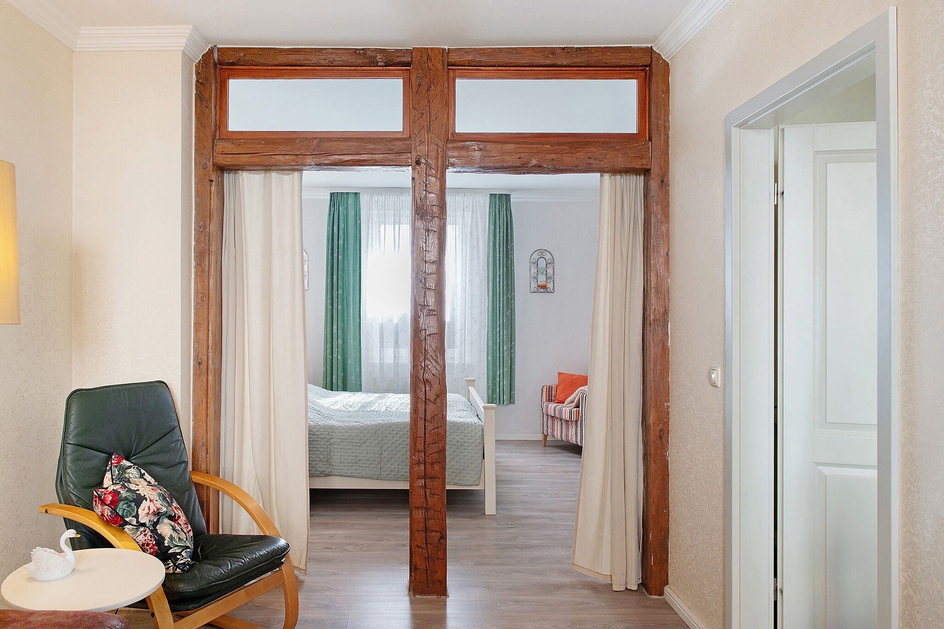 Ein Zimmer mit Holzbalkendecke, einem Bett mit grünen Vorhängen, einem Sessel mit Kissen und einem kleinen Tisch mit einer Schwan-Dekoration.