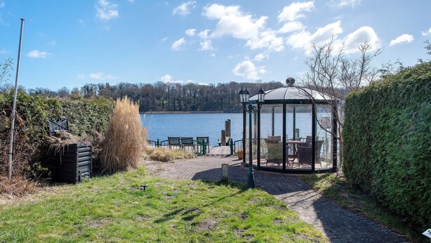 A gazebo with a glass enclosure overlooking a lake, surrounded by a grassy area and a hedge. - Apartment 'Fewo Schwan' with Wi-Fi (Malchow)