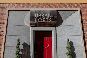 Eine Gebäudefassade mit einem Schild, auf dem "URBAN LOFTS" steht, über einer roten Tür, flankiert von zwei kleinen Bäumen.