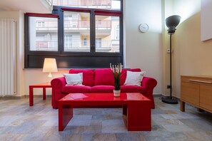 A red sofa with white cushions, a red coffee table, a floor lamp, and a view of balconies through a window. - Loft Vermouth by Wonderful Italy (Torino)