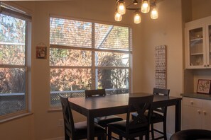 Un coin repas avec une table et des chaises, de grandes fenêtres équipées de stores et un meuble de cuisine.
