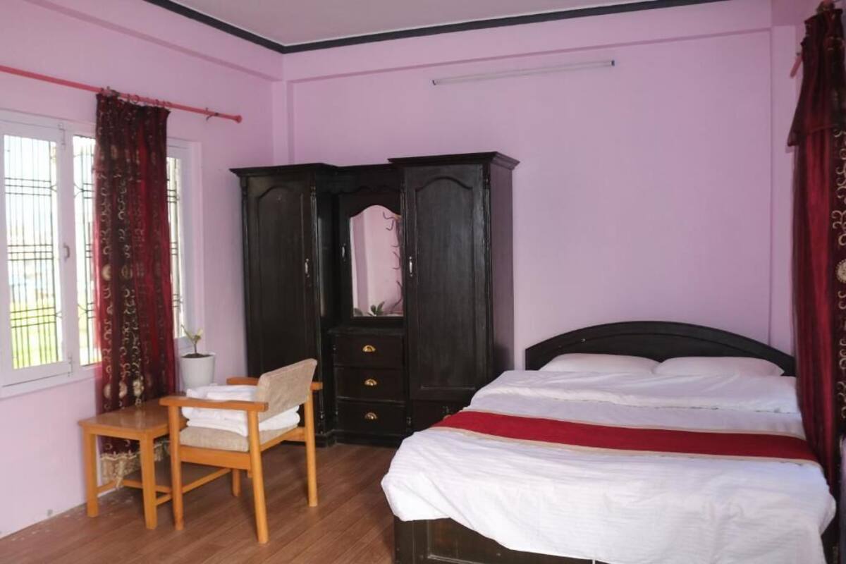 A bedroom with a bed, a wooden chair, a dark wooden wardrobe, and a window with curtains.