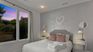Ein Schlafzimmer mit großem Fenster, einem Bett mit weißer Bettwäsche, zwei Nachttischen mit Lampen und einem dekorativen Herz an der Wand.