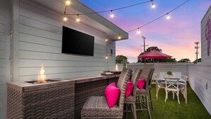 Eine Terrasse im Freien mit Bar, Korbstühle mit rosa Kissen, einem flachbildfernseher an der Wand und Lichterketten.