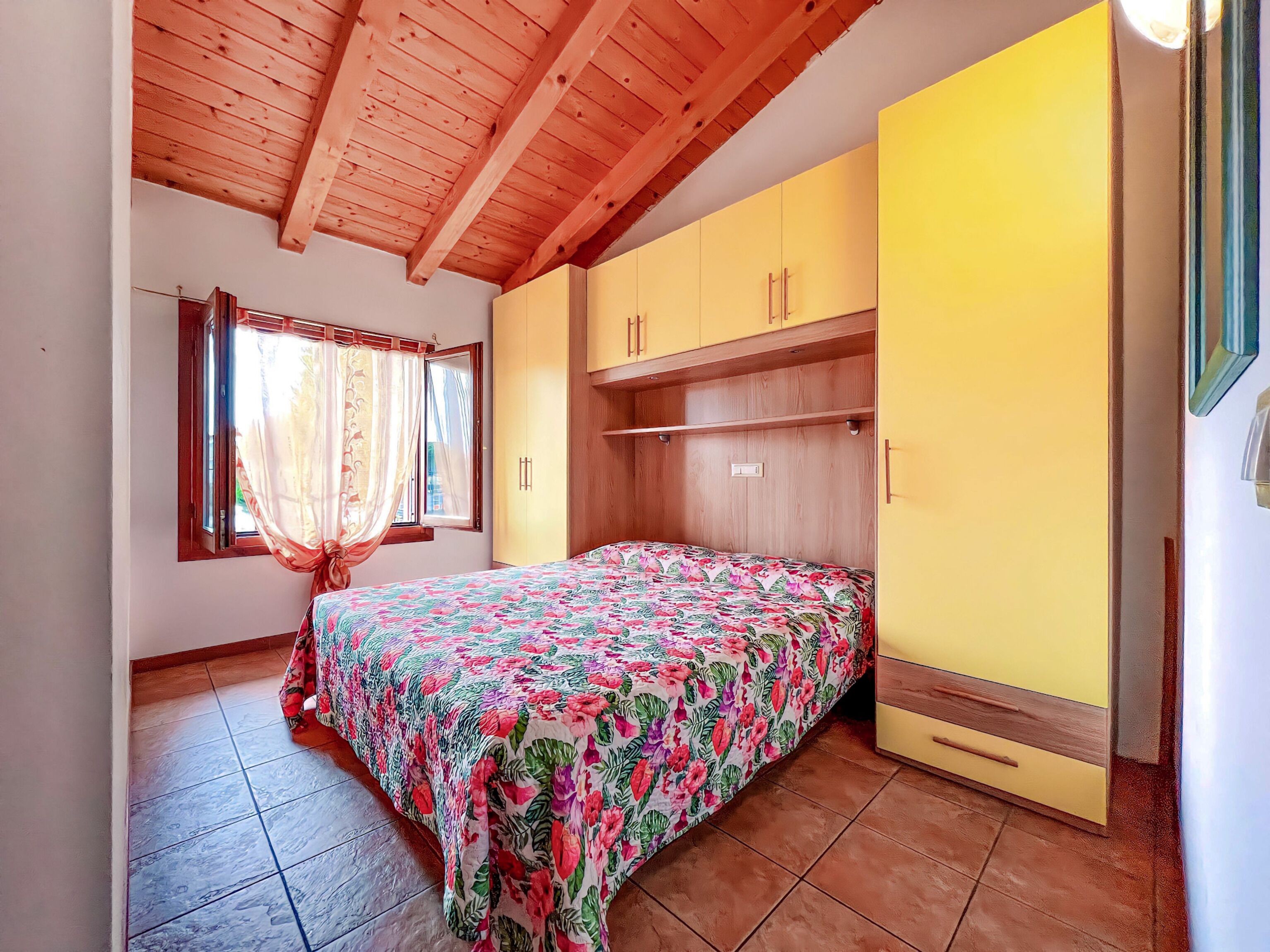 A bedroom with a wooden ceiling, a bed with a floral bedspread, a window with curtains, and a yellow wardrobe.
