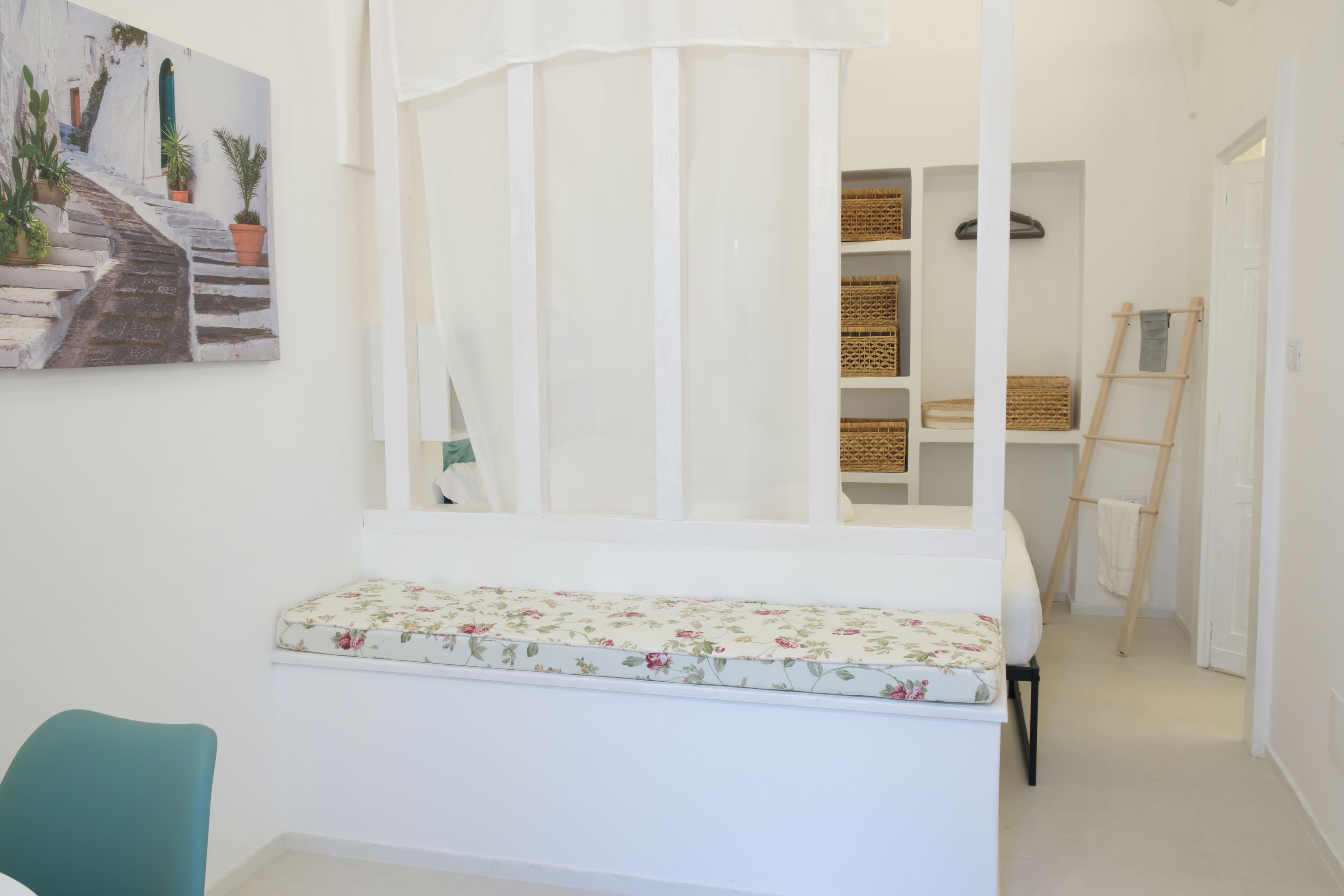 A room with a bed, a window with a white curtain, a painting on the wall, and a ladder leaning against the wall.